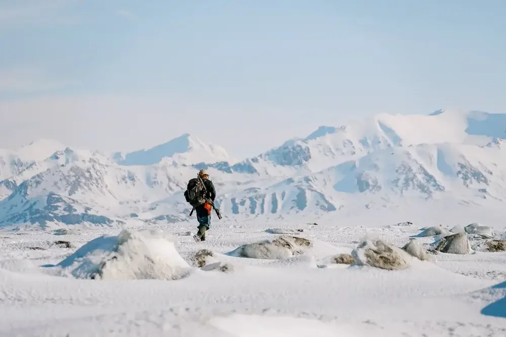 A person dressed in winter gear carrying a backpack and walking across a snowy landscape with mountains in the background.
