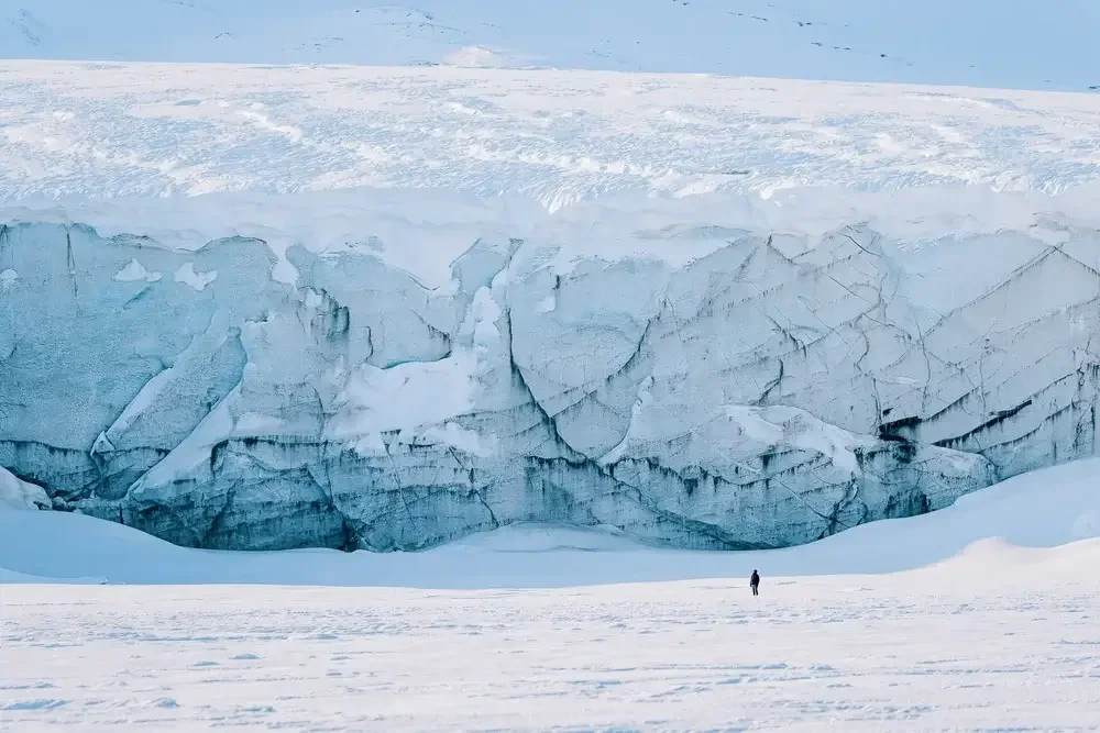 A person standing on snow in front of a large ice cliff in a cold, icy landscape.

