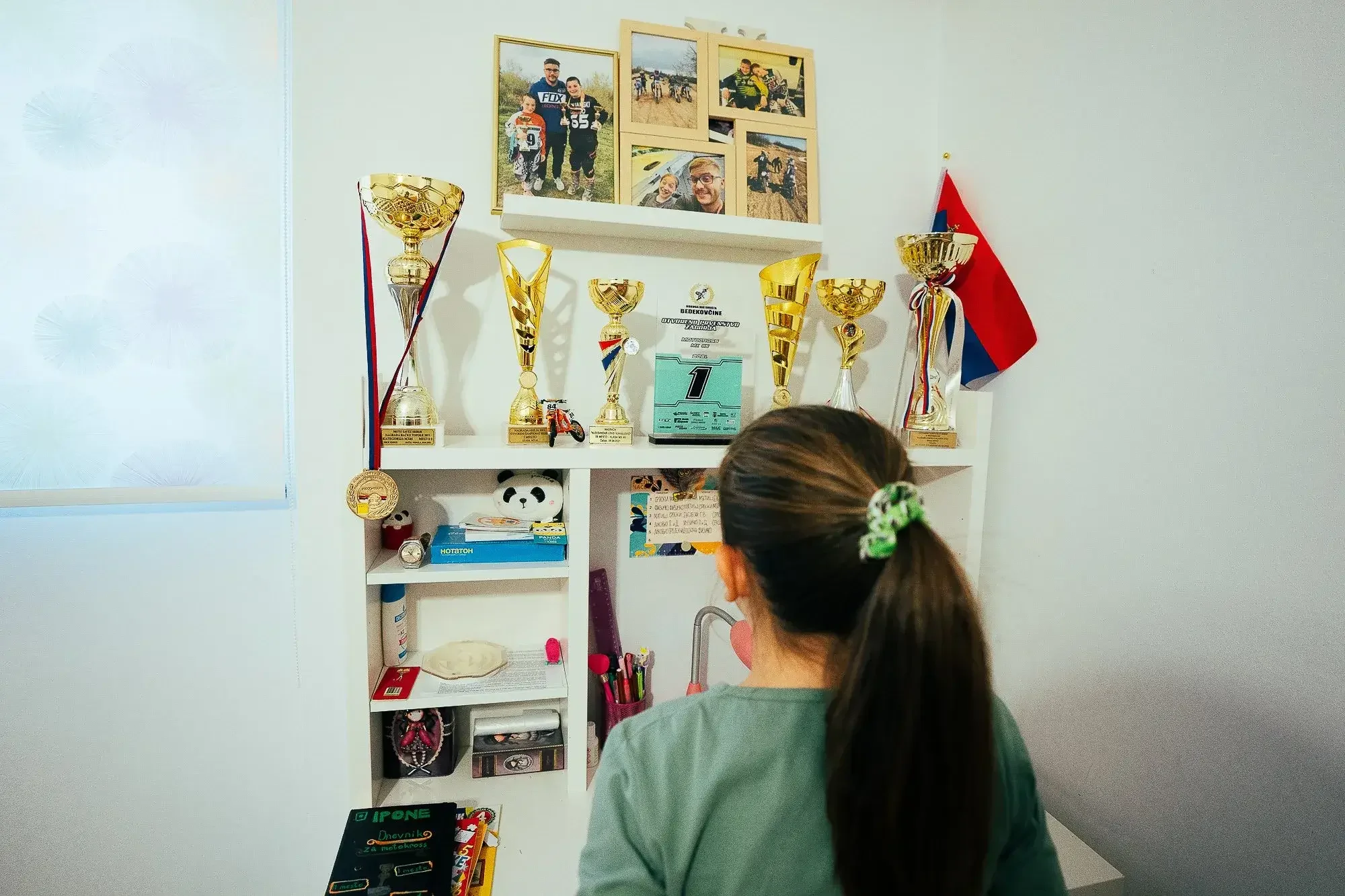 Girl with long hair tied back, wearing a green top, looking at a white shelf with trophies, medals, and framed family photos above the shelf. A red and blue flag is on the right.
