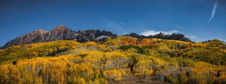 A mountain landscape with colorful autumn trees in shades of yellow, orange, and green, under a blue sky with wispy clouds.

