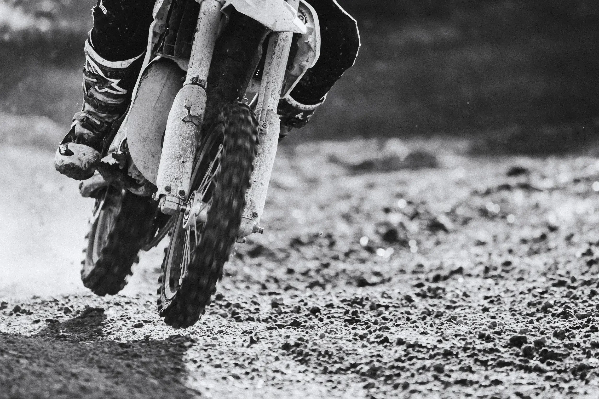 Close-up of a dirt bike racing on a dirt track, captured in black and white from a low angle.
