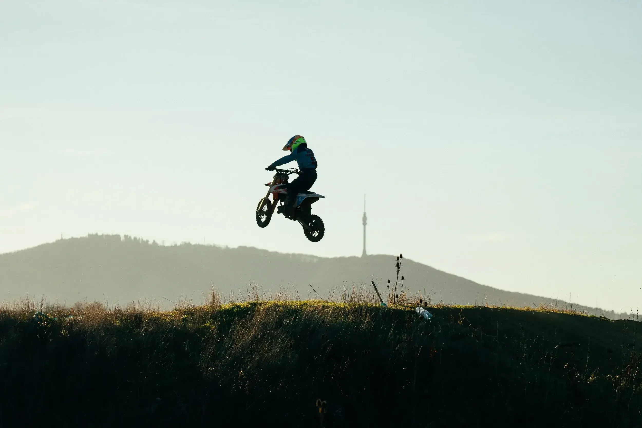 A person riding a dirt bike in mid-air during a jump on a natural outdoor trail with hills and a distant tower in the background.
