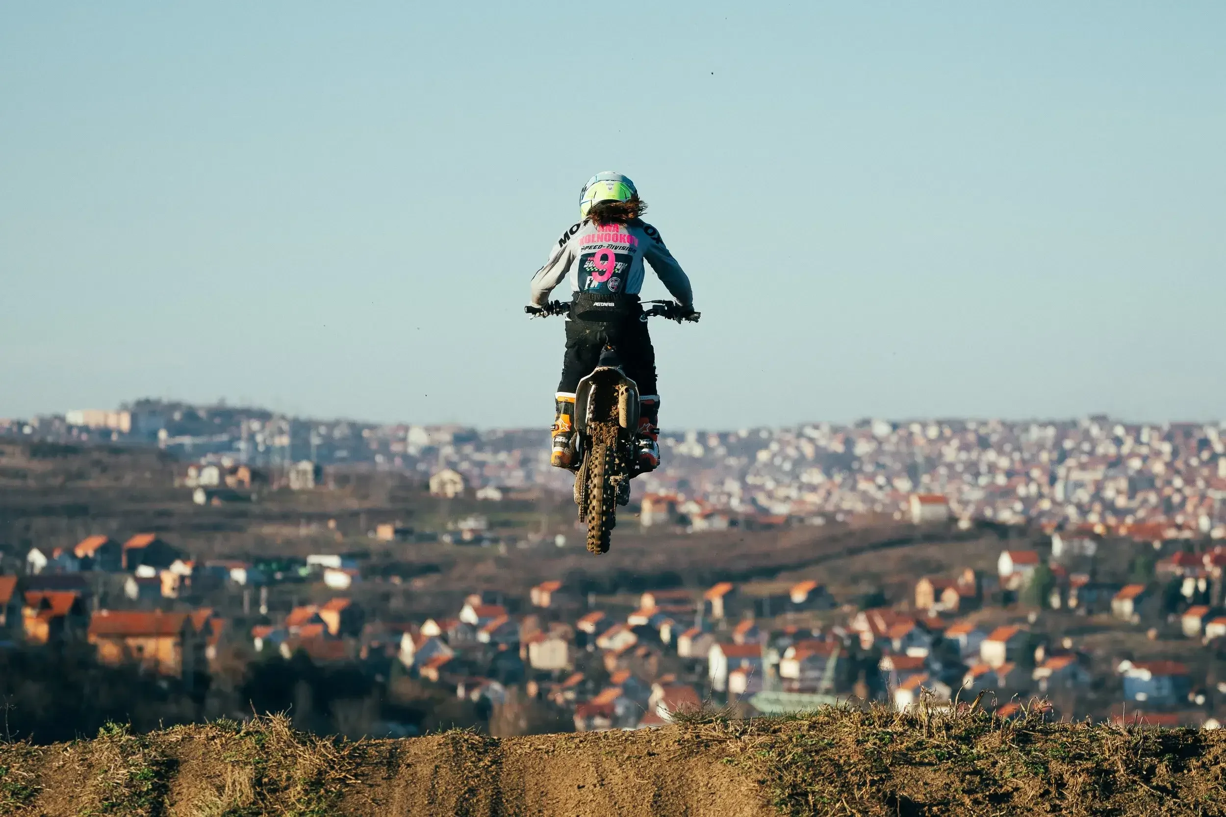 A person riding a dirt bike jumps into the air over a dirt track with a town in the background.

