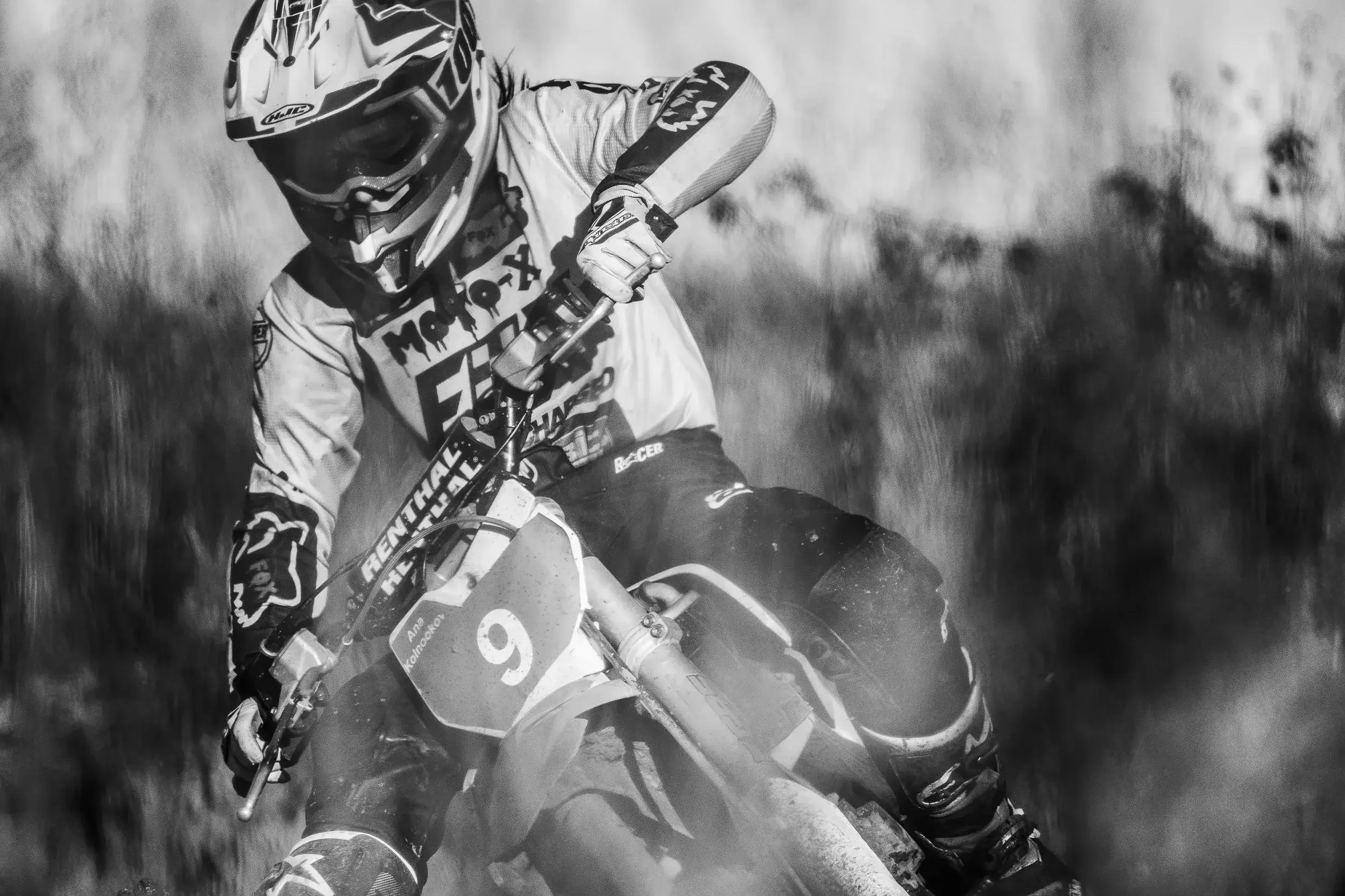A black and white photo of a motocross rider leaning sharply into a turn on a dirt track, wearing a helmet and protective gear, with dust flying around.

