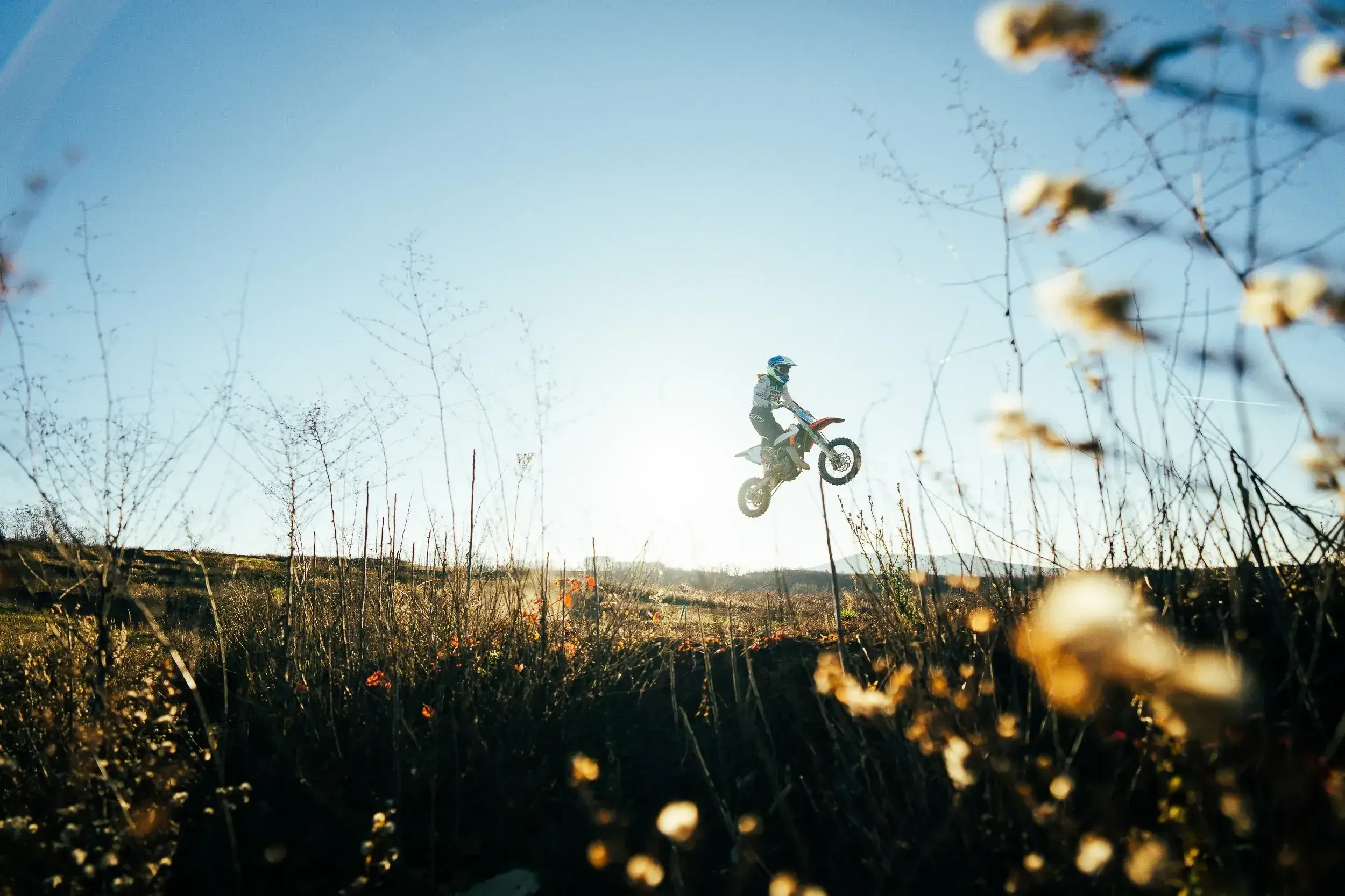 A person riding a dirt bike airborne during a jump in an outdoor setting with dry grass and bushes under a clear blue sky.

