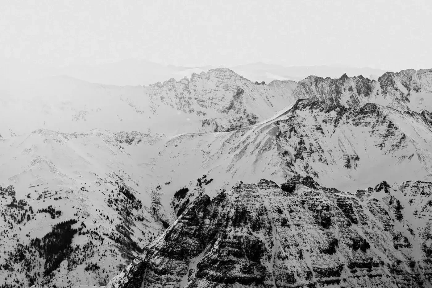 Black and white photograph of snow-covered mountain peaks and ridges, with some forests visible in the lower part of the image.