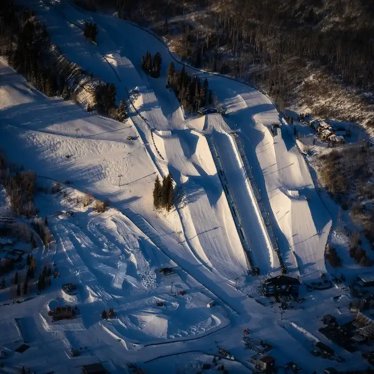 An aerial view of a ski jump ramp covered in snow surrounded by trees and buildings in a winter landscape.
