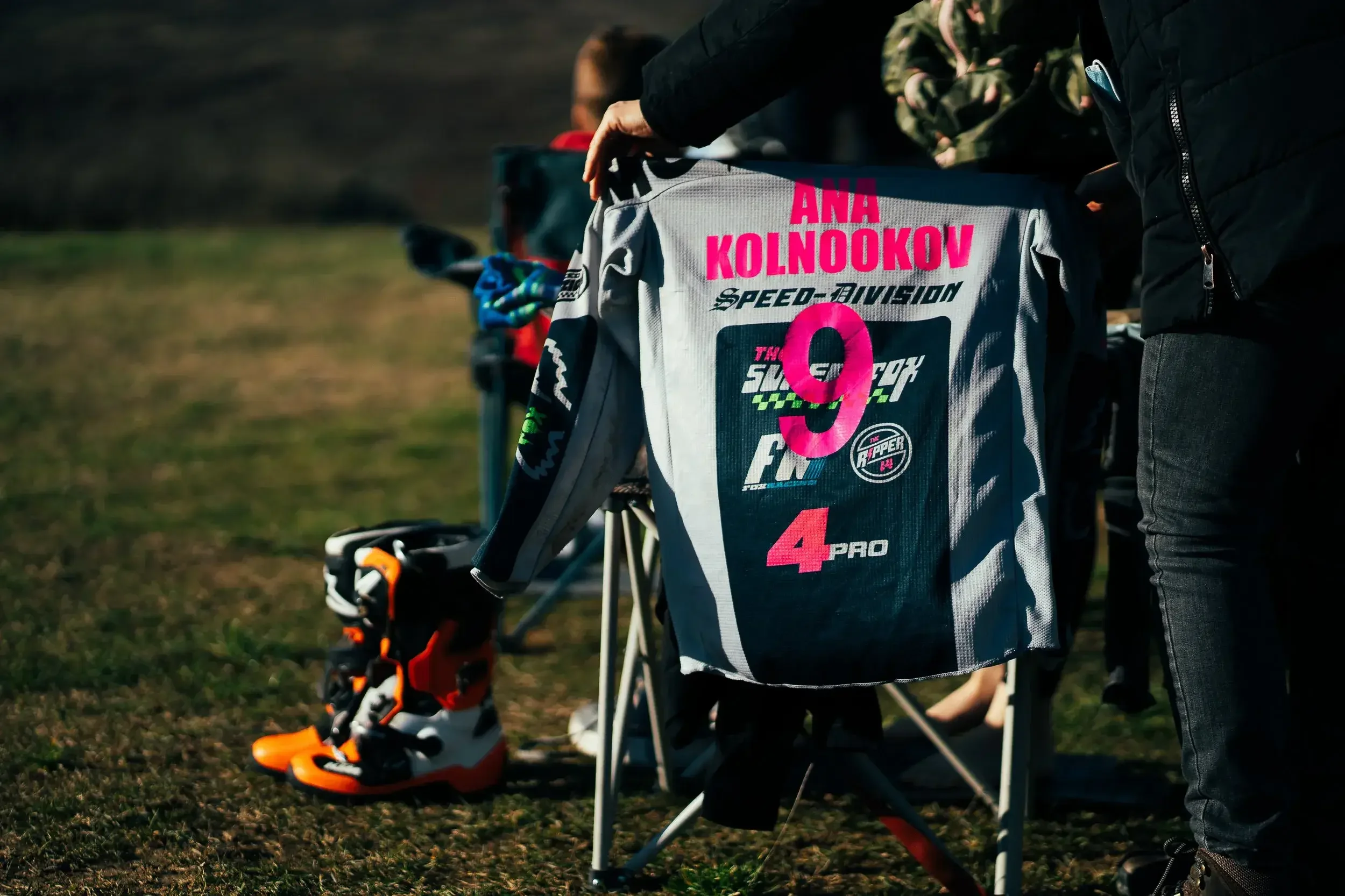 A gray and pink motocross racing suit with the name 'ANA KOLNOOKOV' hanging on a chair, with motocross boots on the ground nearby, outdoors on grass.

