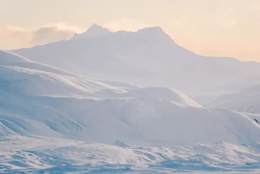 Snow-covered mountain landscape with rolling snow fields and distant peaks under a pale sky.
