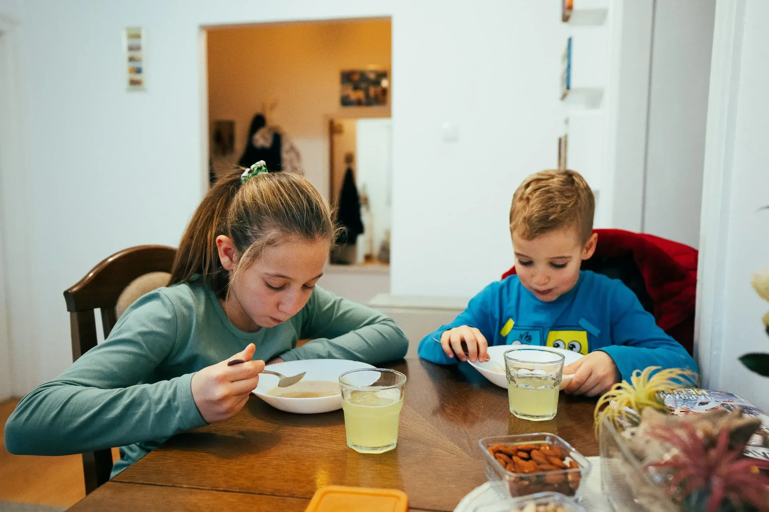 A girl and a boy sitting at a dining table, eating. The girl has long brown hair in a ponytail, and the boy has short red hair. There are glasses of yellow juice and bowls of food on the table.

