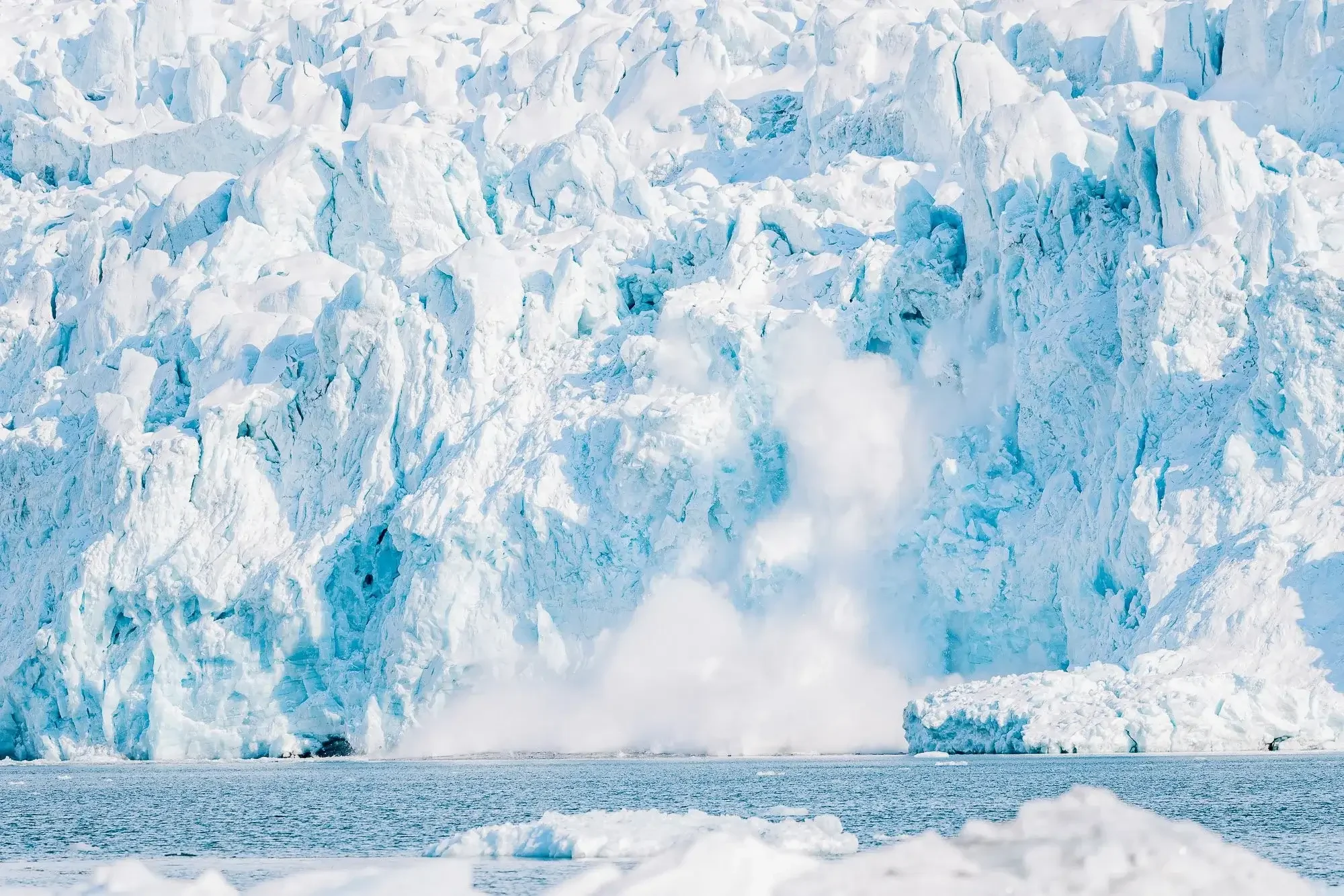 Large glacier calving into the ocean, with chunks of ice breaking off and crashing into the water.