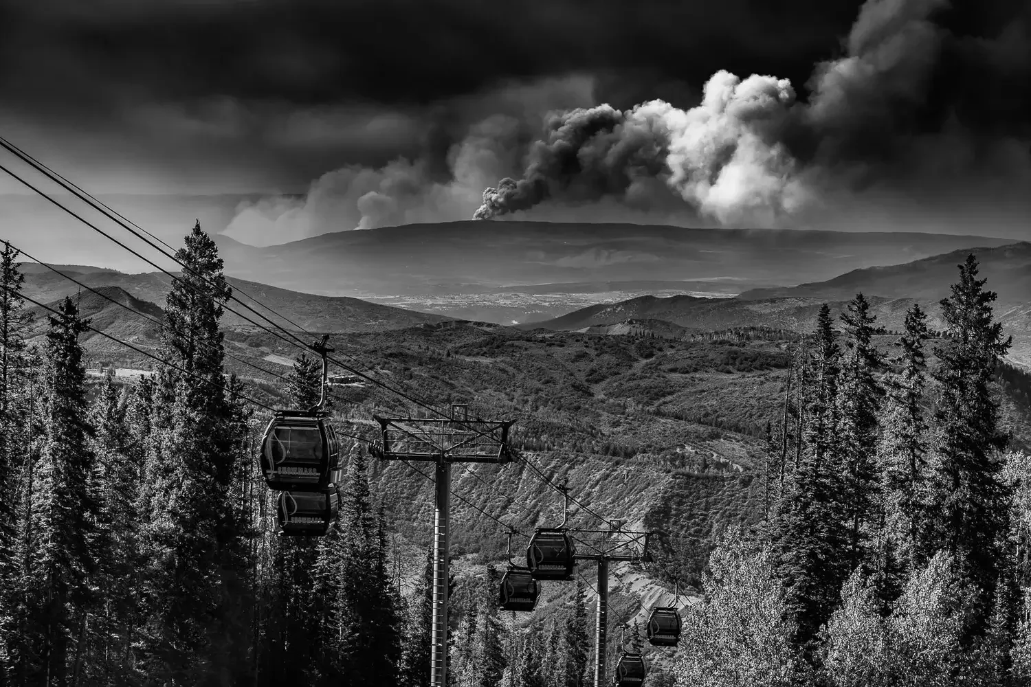 Black and white photo of a ski lift ascending through a forested mountain landscape with smoke and ash rising from distant volcanic activity on the horizon.