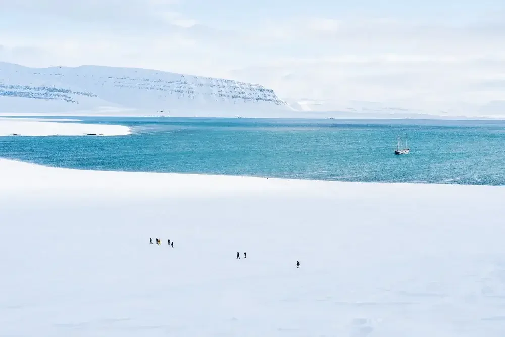 A snowy landscape with small groups of people walking on the snow, a calm blue sea, a sailboat, and snow-covered mountains in the background.
