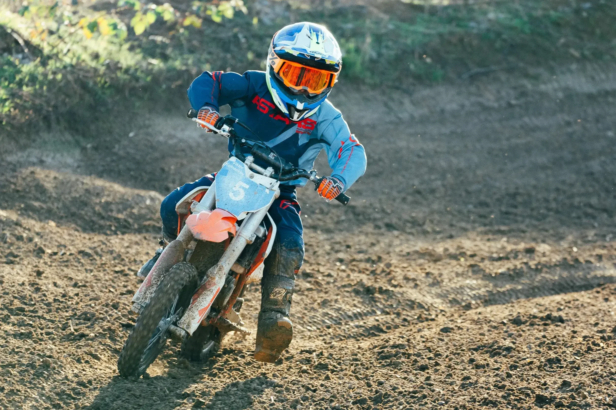 A motocross rider in blue and orange gear riding a dirt bike on a rough dirt track, wearing a helmet with orange goggles.
