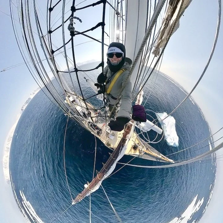 Tamara Susa taking a selfie high above the ocean aboard a sailing ship with sails and rigging, with a circular fisheye effect.