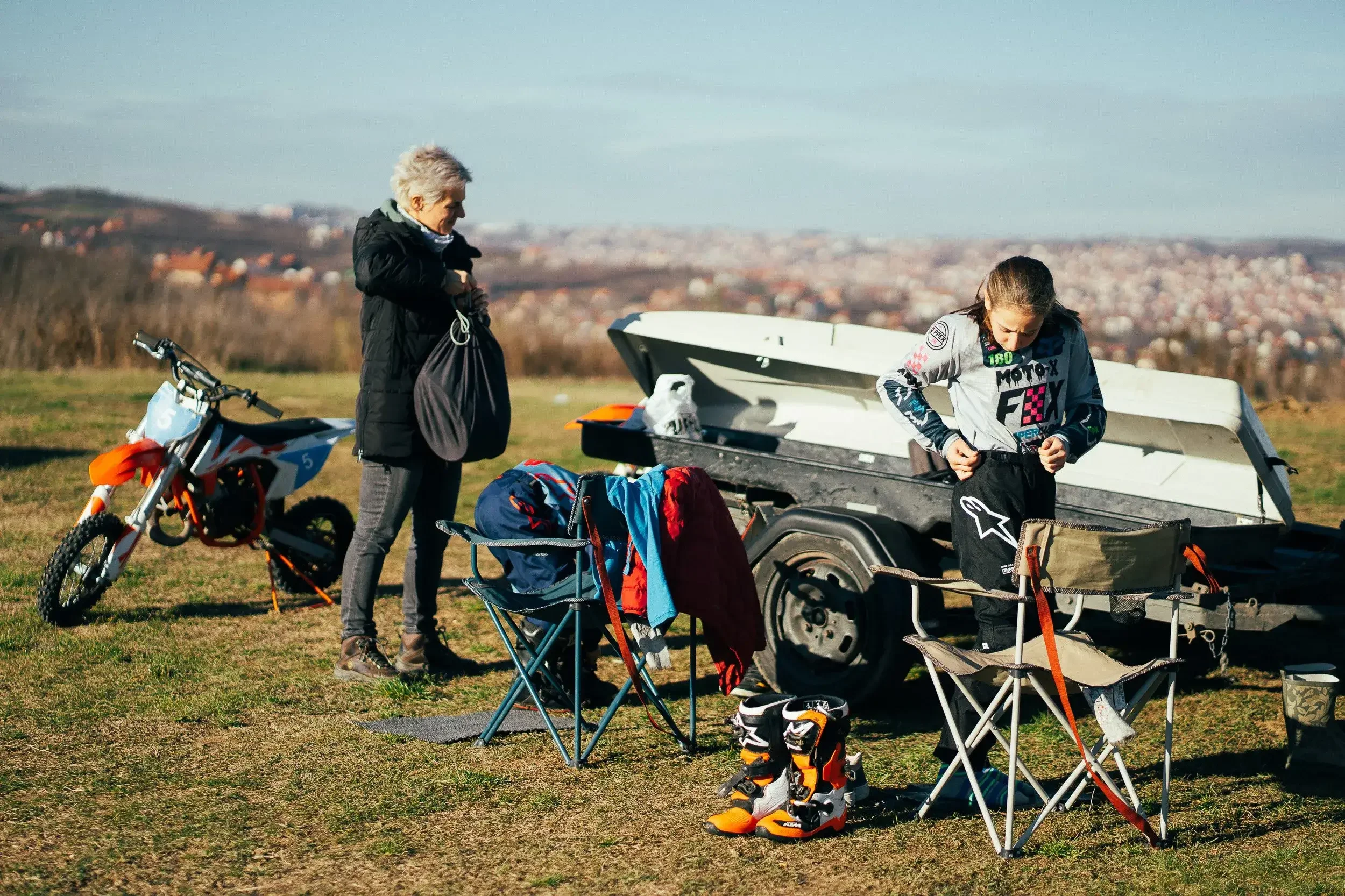 Two women preparing for motocross riding outdoors on a grassy field, one standing near a dirt bike and the other adjusting clothing near a trailer with motocross gear.
