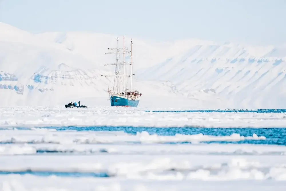 A blue sailing ship and a smaller boat with people sailing through icy waters surrounded by snow-covered mountains and ice floes.
