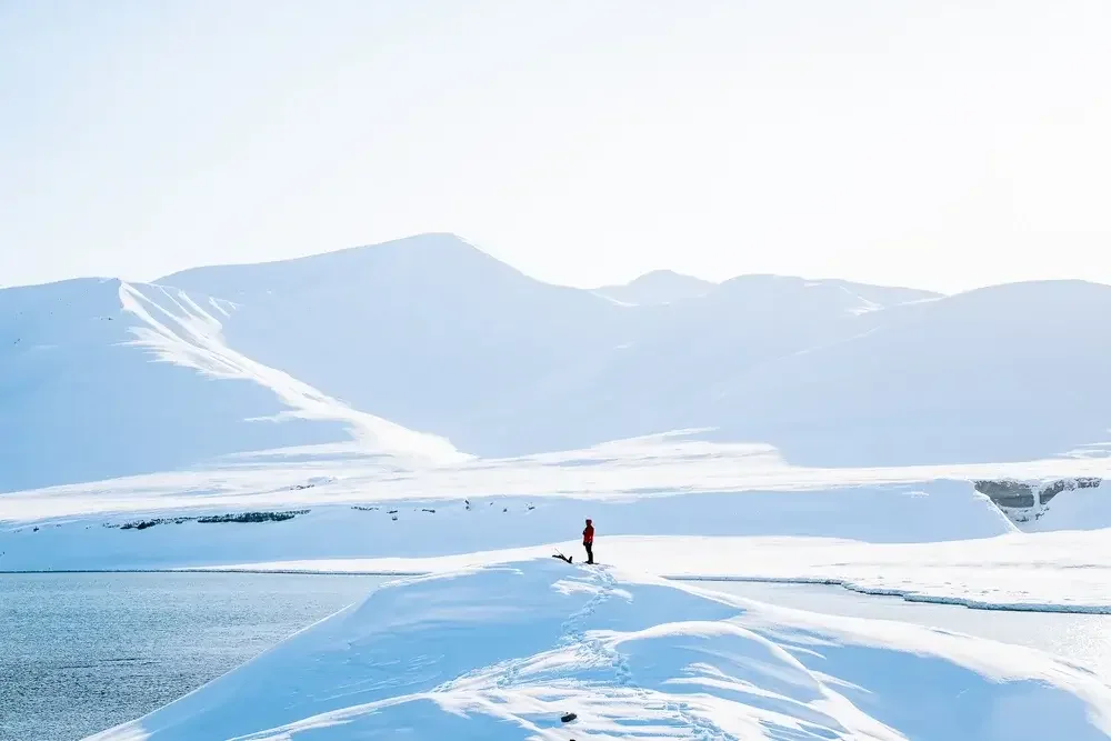 A person in red outdoor clothing standing on snow-covered terrain near a water body in a cold, mountainous landscape with snow-covered peaks in the background.
