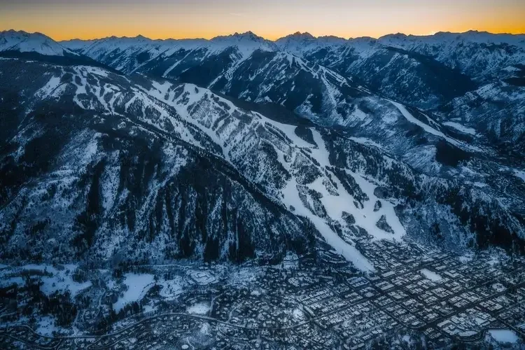 An aerial view of a snow-covered mountain range at sunset, with a town at the base of the mountains.
