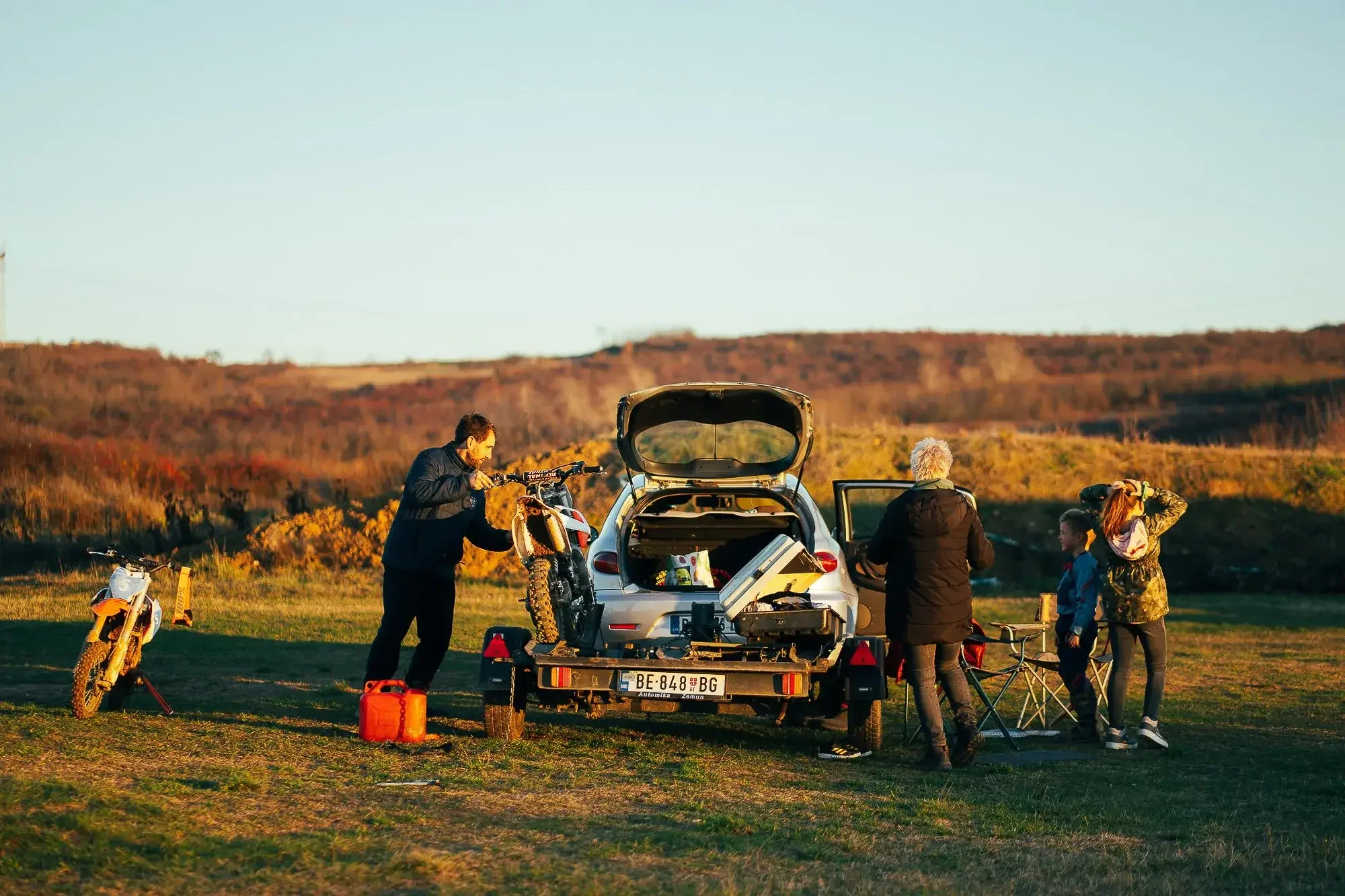 Group of people preparing for outdoor activities with bikes, car, and camping gear in a grassy field during sunset
