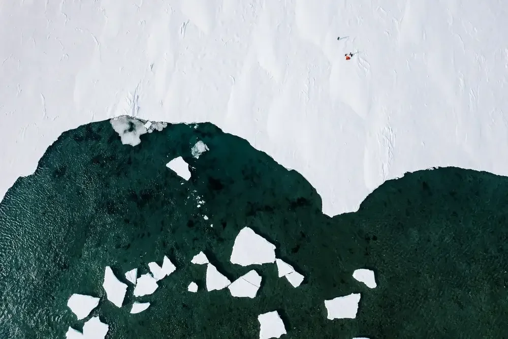Aerial view of an icy coastline with floating ice chunks and dark blue ocean water.
