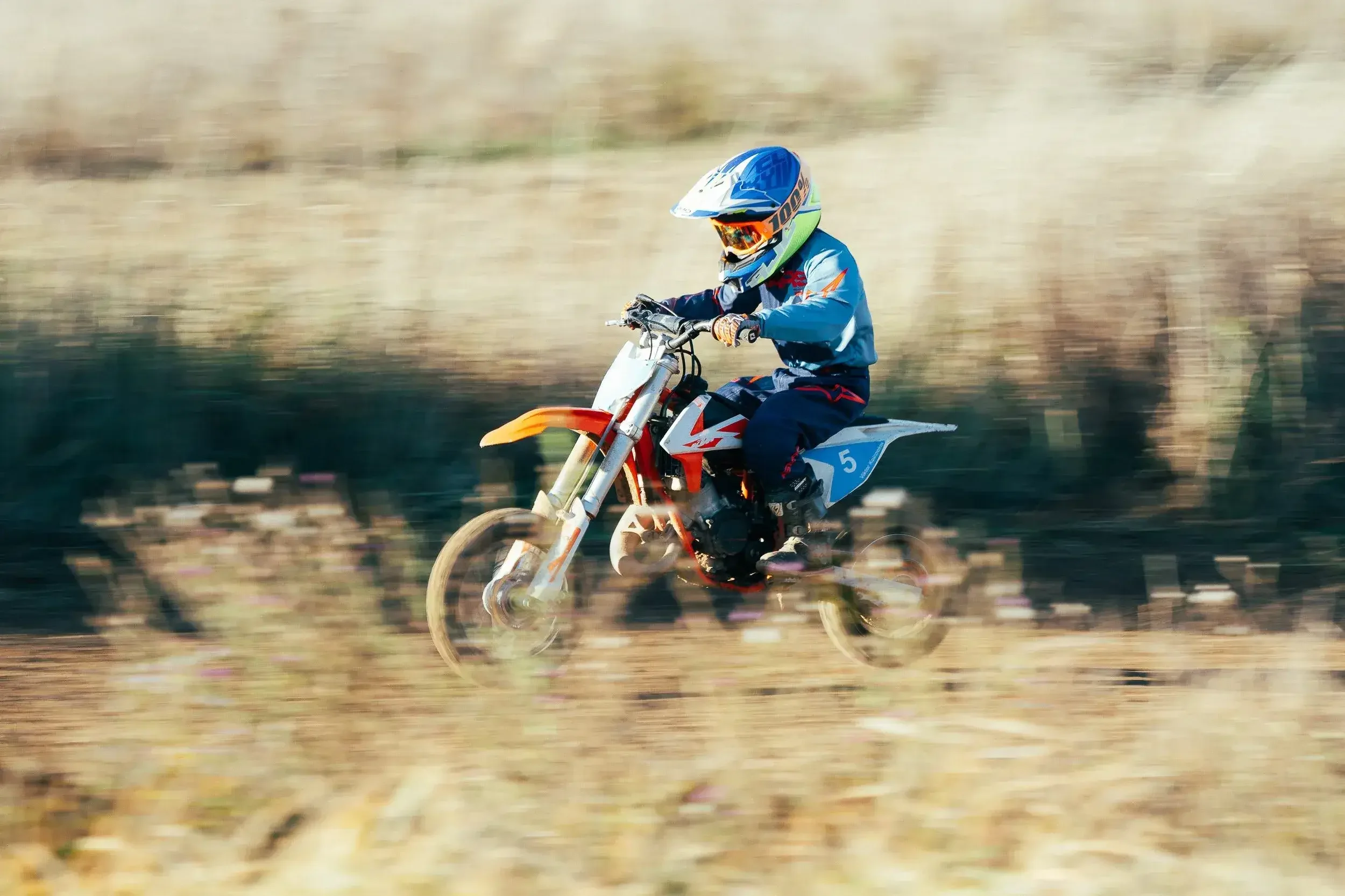 A person in motocross gear riding a dirt bike on a trail, with blurred background indicating motion.
