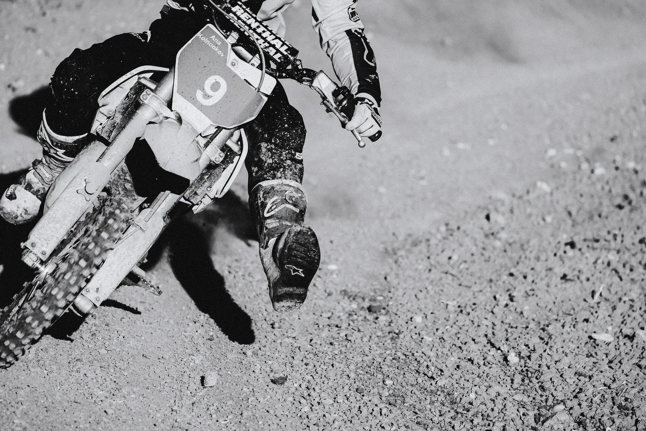 A motocross rider wearing protective gear is riding a dirt bike on a gravel surface, leaning into a turn.
