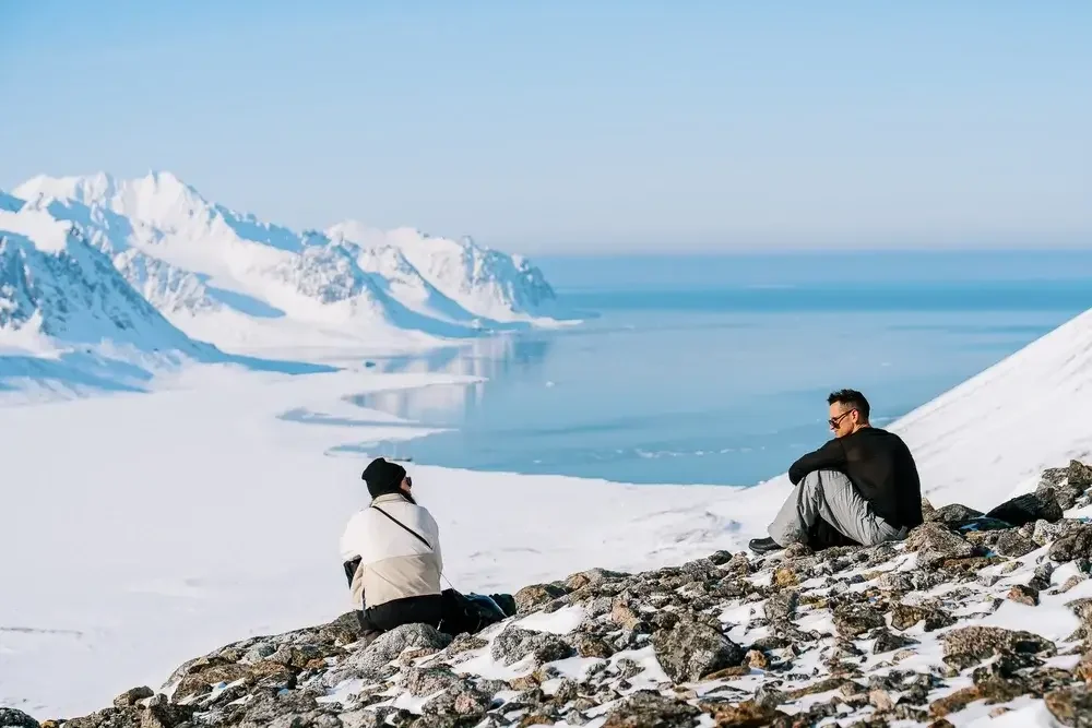 Two people sitting on rocky snowy terrain overlooking an icy ocean with snow-covered mountains in the background.
