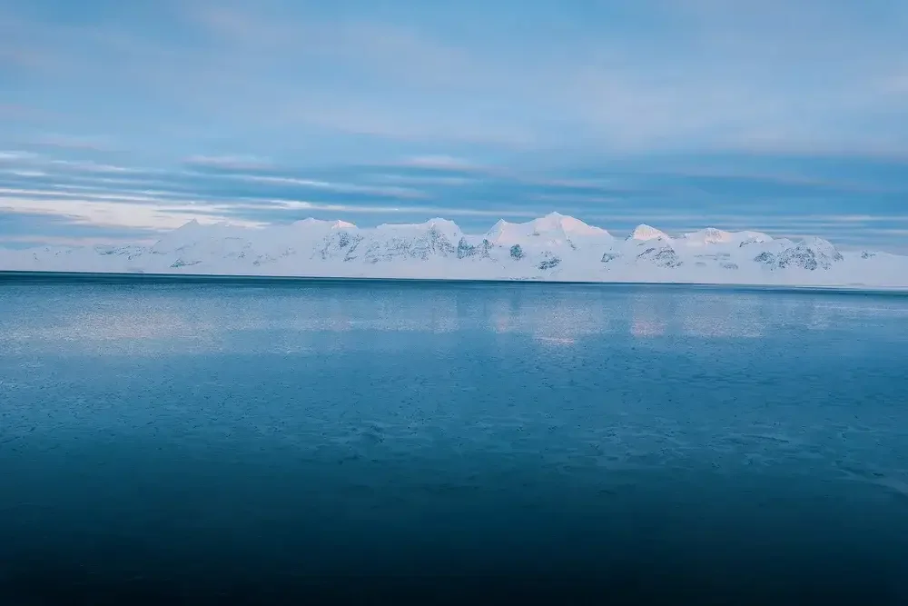 Snow-covered mountains across calm blue waters under a cloudy sky.
