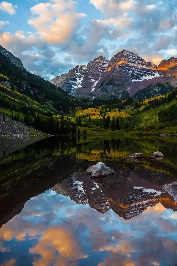 Mountain landscape with snow-capped peaks, green forests, and a calm lake reflecting the mountains and sky, during sunset or sunrise.
