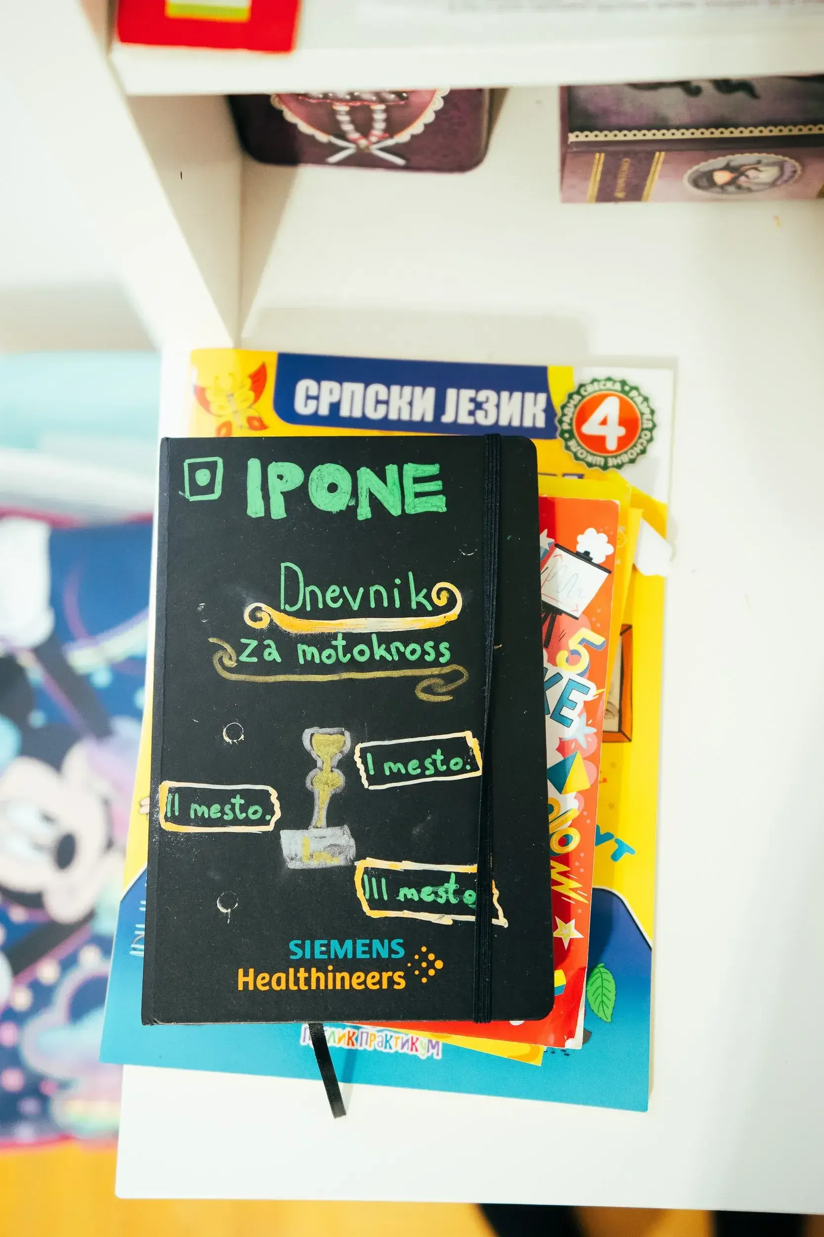 A black notebook with colorful writing and drawings on the cover, placed on top of colorful children's books on a white shelf.
