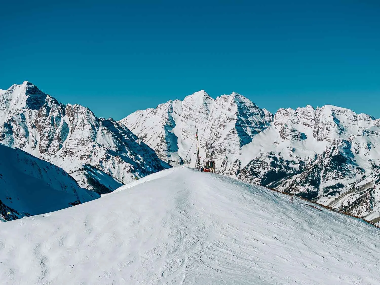 Snow-covered mountain landscape with clear blue sky, featuring a ski slope, ski lift, and rugged peaks in the background.