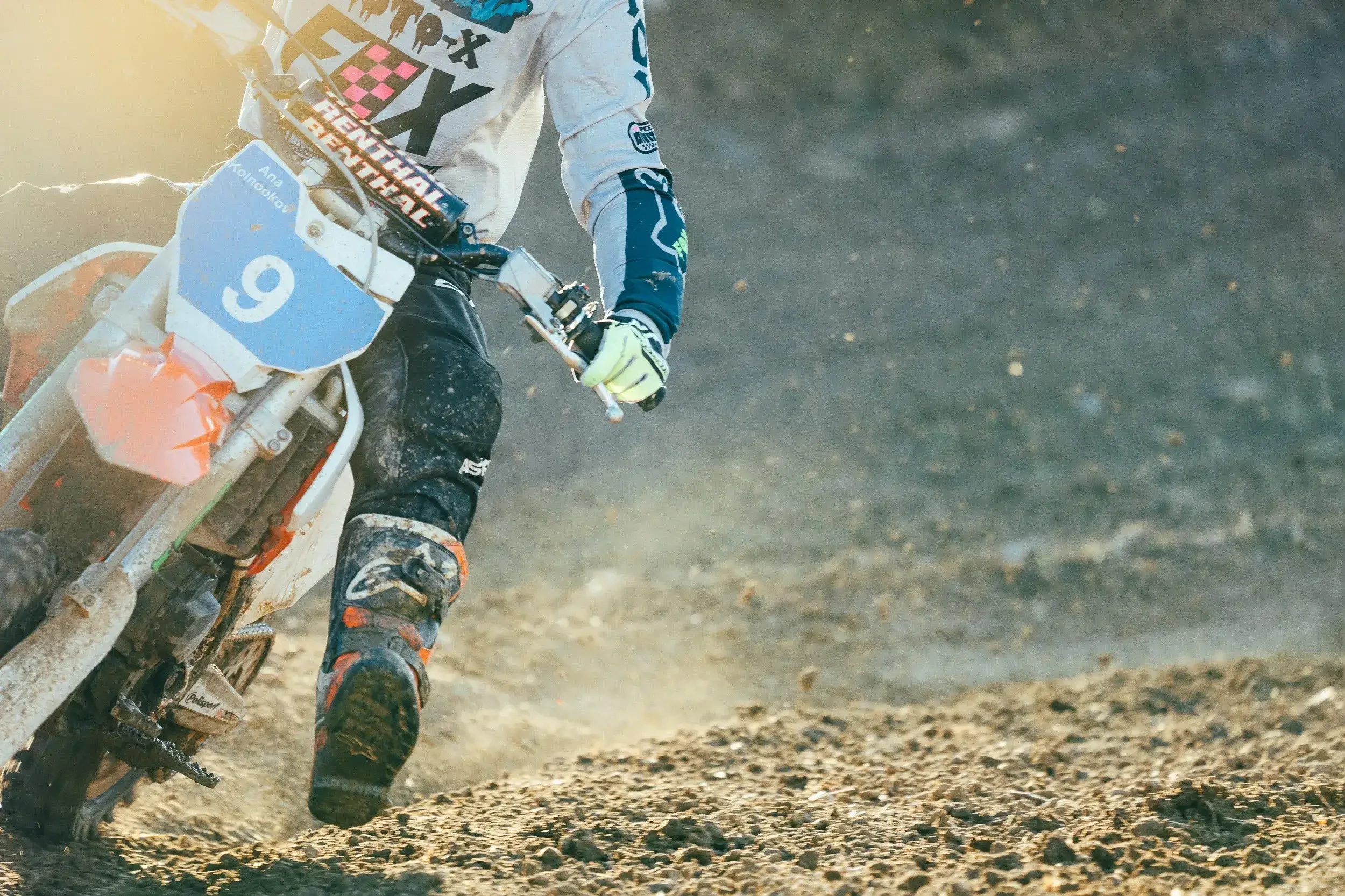 Motocross rider wearing protective gear riding on a dirt track with dust and gravel in the air.
