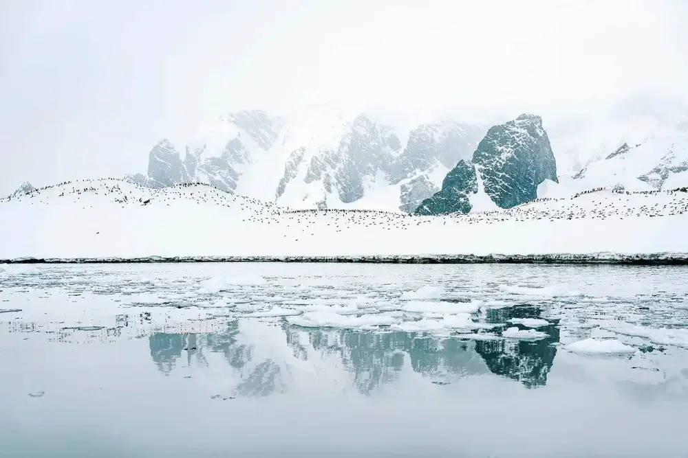 Snow-covered mountains and rocky peaks reflected in a partially frozen body of water, with a drifting snow or ice on the surface.