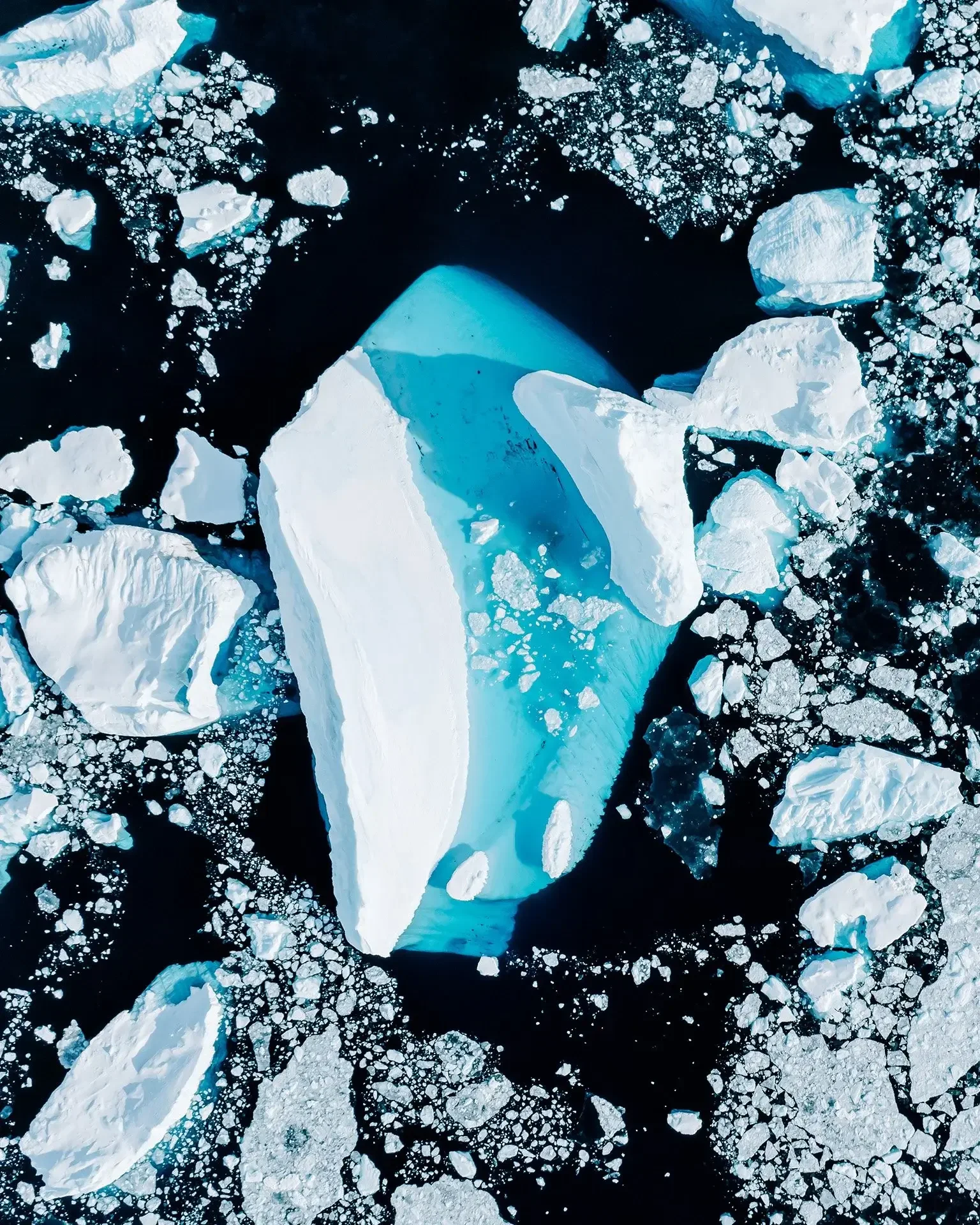 Aerial view of large iceberg floating in dark ocean with smaller ice fragments surrounding it.