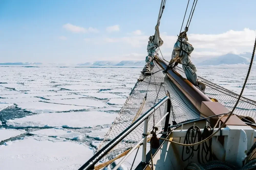 View from a sailboat navigating through icy, frozen waters with cracked ice and snow-covered mountains in the background.
