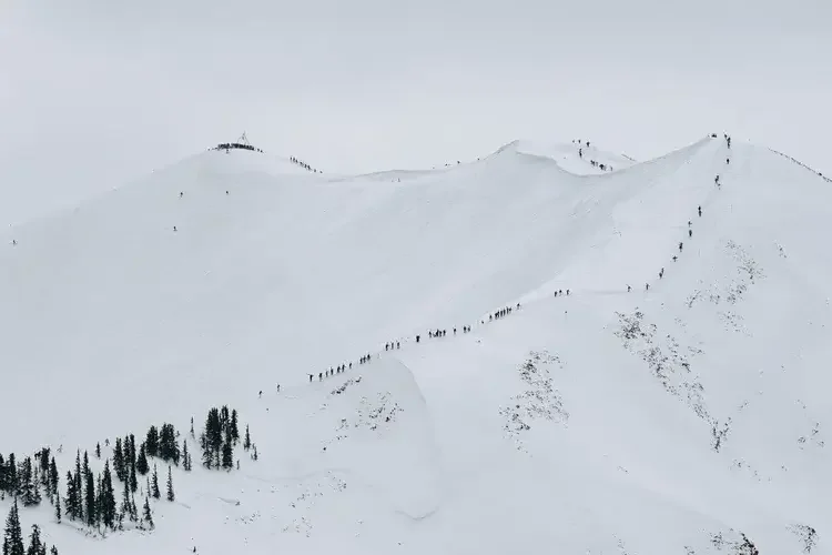 A large group of people hiking up a snowy mountain ridge with some trees at the bottom and a cloudy sky overhead.
