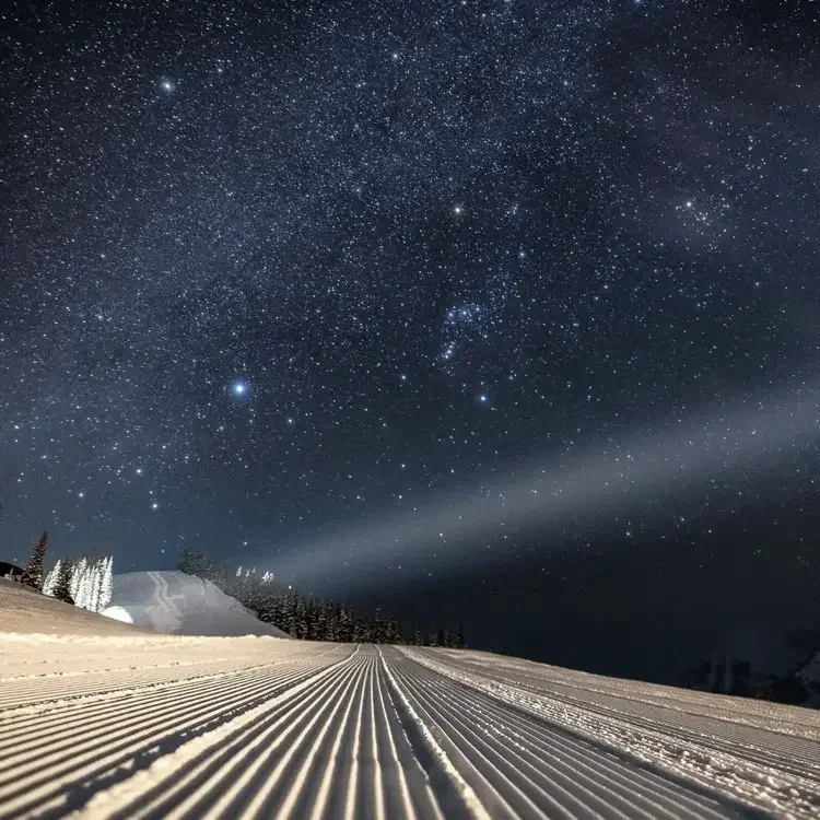 Nighttime snowy landscape with groomed ski trail under a star-filled sky, with some trees and mountains in the background.
