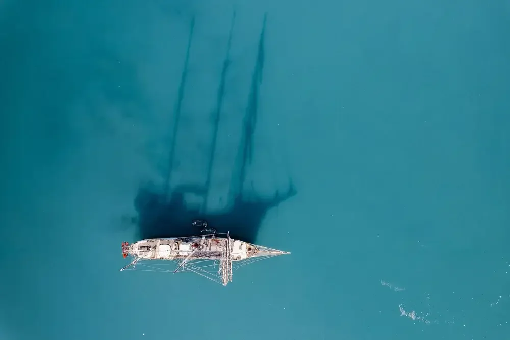 An aerial view of a sailboat on turquoise water with its shadow cast on the surface.
