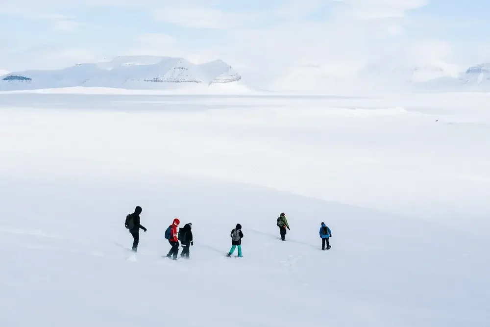 A group of people walking across a snow-covered landscape near a body of water with snow-covered mountains in the background.
