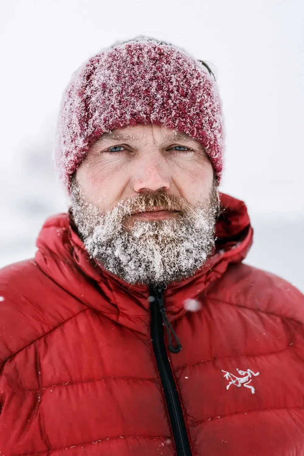 A man with a frost-covered beard, wearing a red beanie and red insulated jacket, standing outdoors in a snowy environment.

