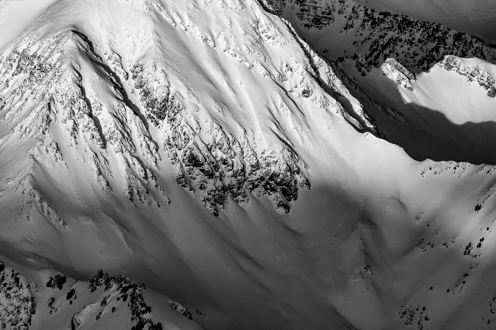 A black and white aerial view of steep snow-covered mountains with rugged rocky outcroppings.