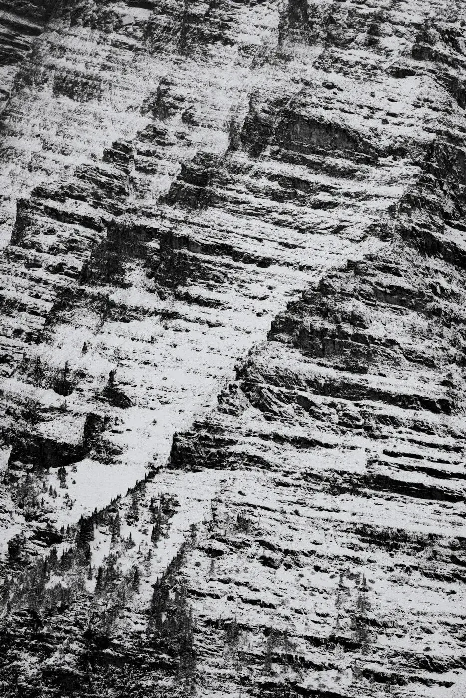 Snow-covered rugged mountain with steep, layered rock formations and sparse trees at the base.
