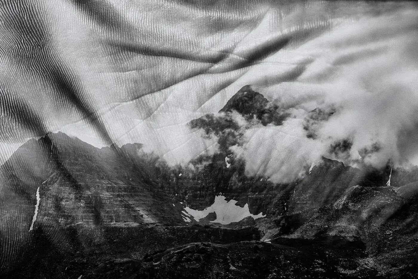 Black and white photo of a mountain landscape with clouds and snow patches, overlaid with a semi-transparent image of textured fabric or netting.
