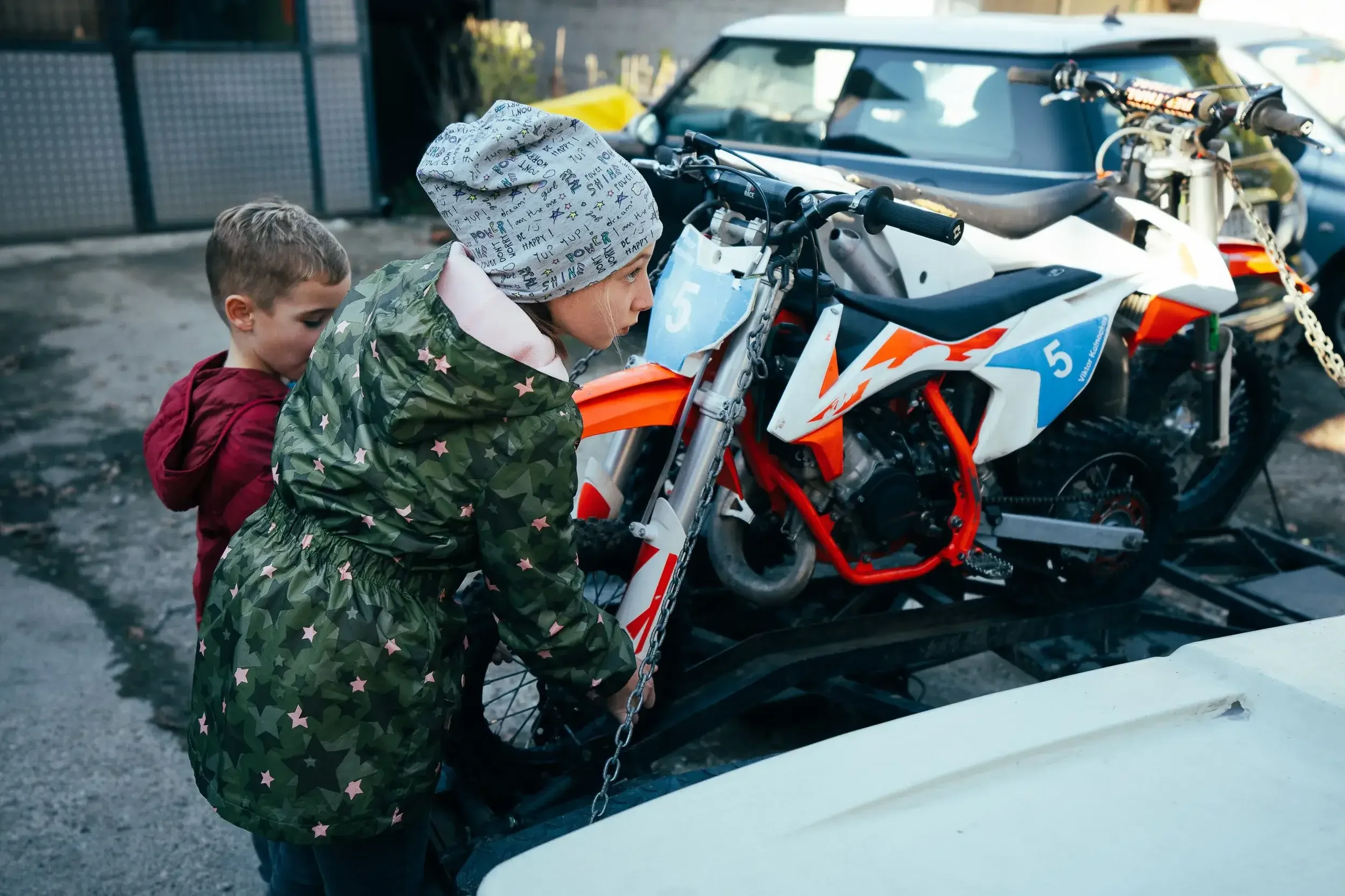 Two children, a girl wearing a beanie and a green star-patterned jacket and a boy in a red jacket, are looking at a motocross bike on a trailer in an outdoor setting.
