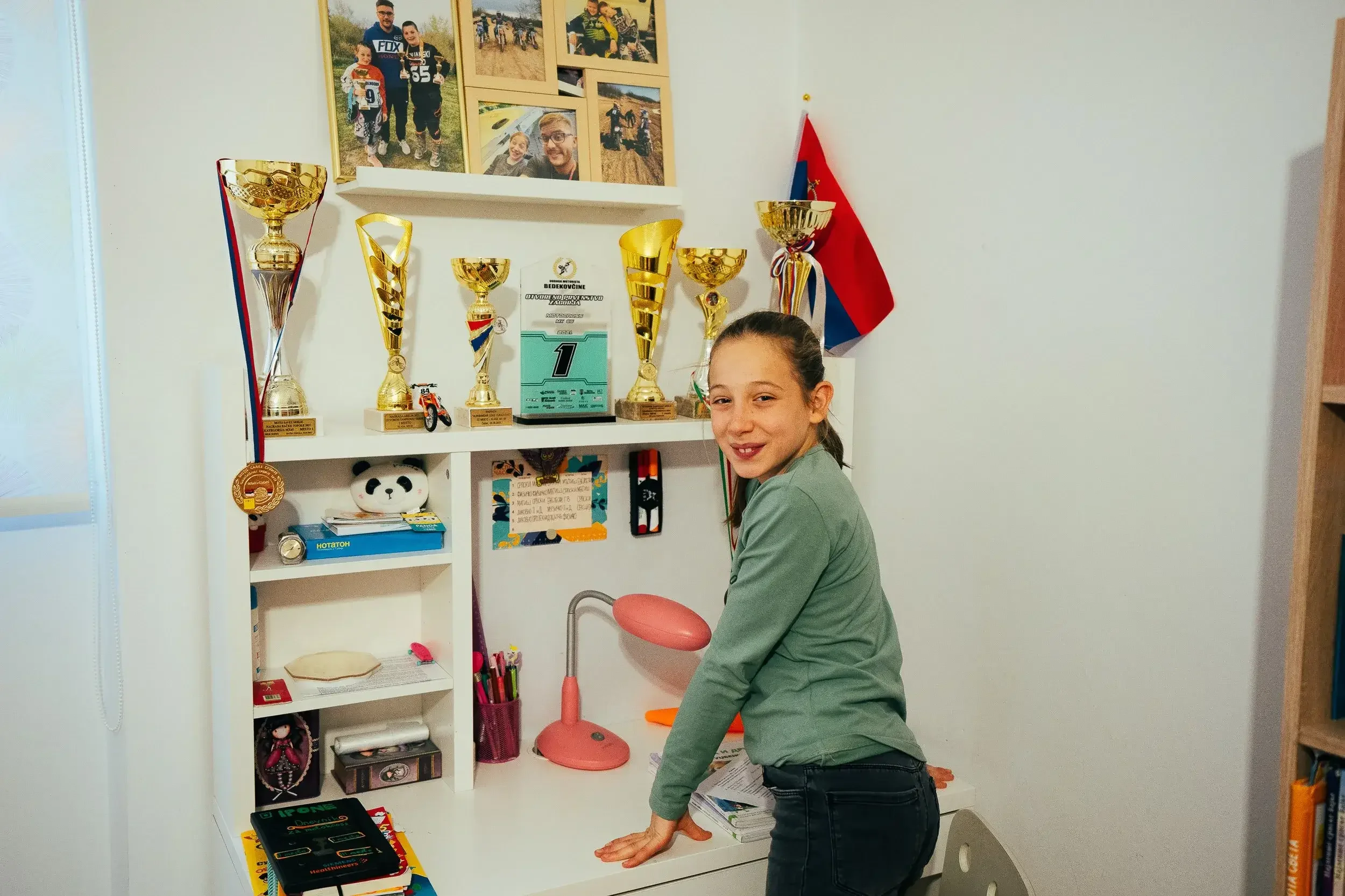 A young girl standing at a white desk with her hands on the surface, smiling at the camera. Behind her is a shelf displaying multiple trophies, a backpack, and other personal items. Above the shelf, there are framed photos and a flag on the wall.