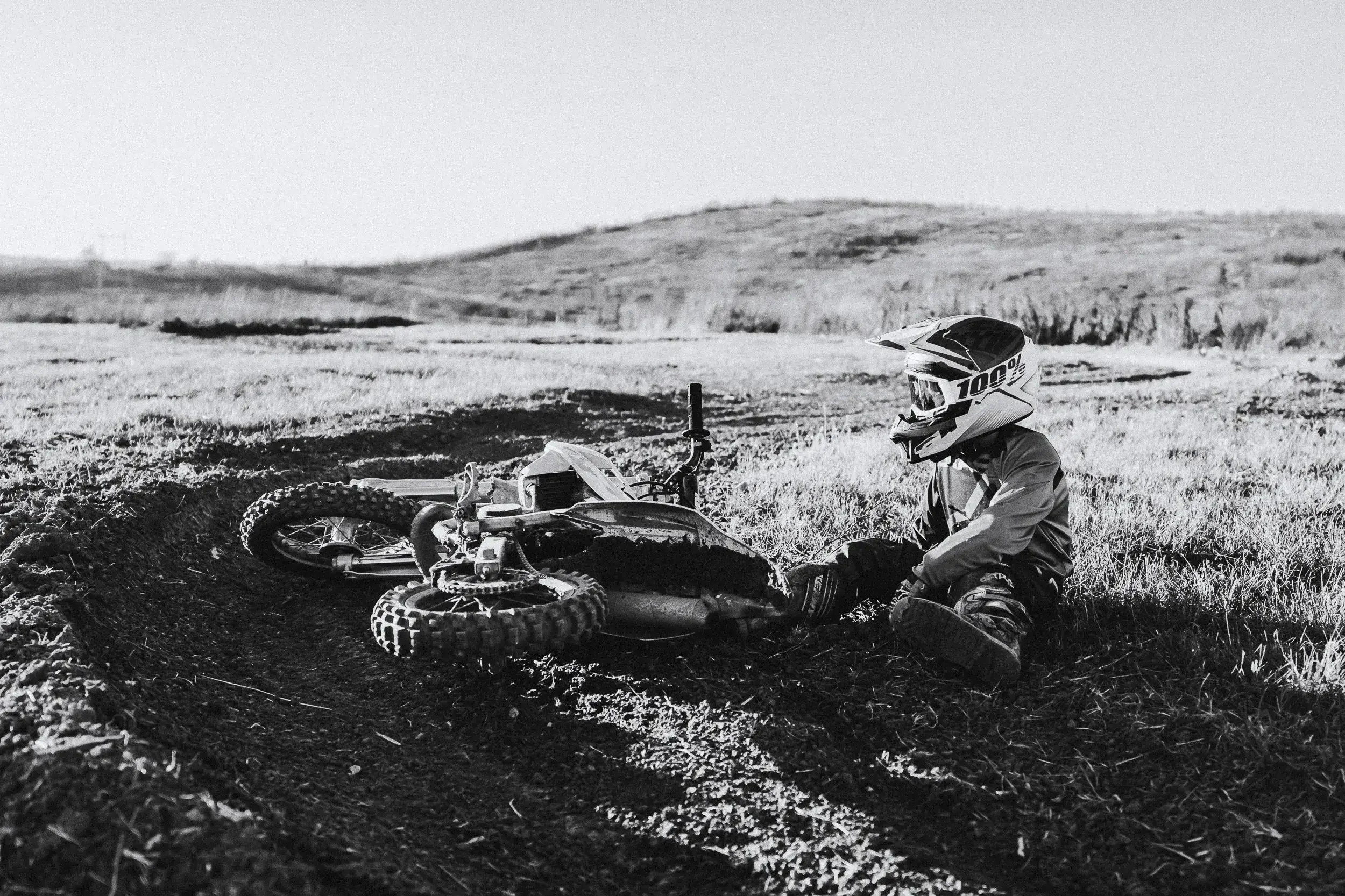 A person sitting on the ground wearing a helmet and motocross gear next to a fallen dirt bike on a dirt trail in an open field.
