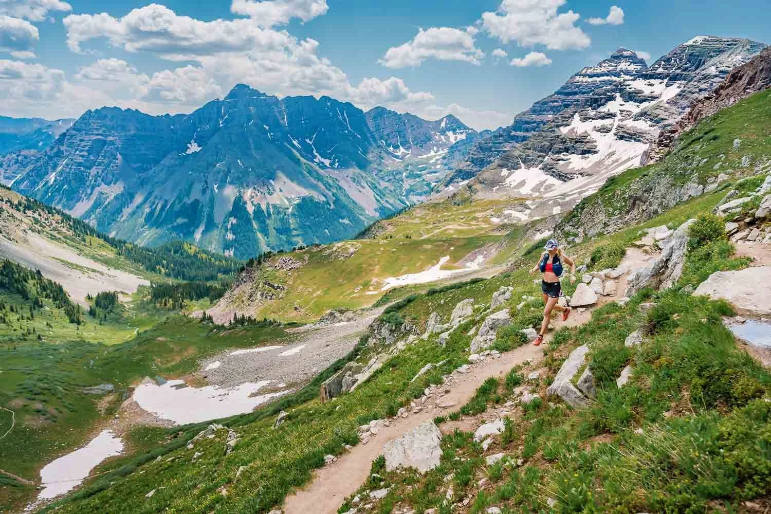 A woman trail running on a narrow mountain trail through a lush green valley with snow patches, surrounded by towering snow-capped mountains and a partly cloudy sky.
