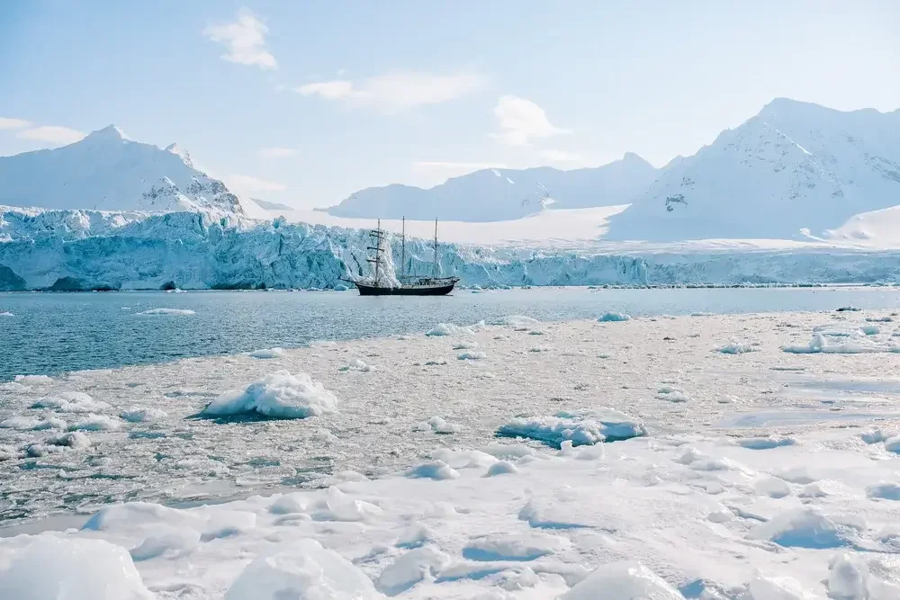 A snowy, icy landscape with a ship sailing near a large glacier and snow-covered mountains in the background.