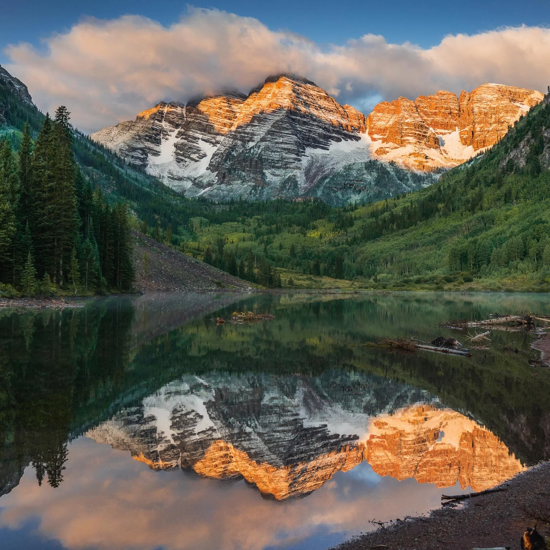 Snow-capped mountains reflected in a calm lake with green forested slopes at sunrise or sunset.